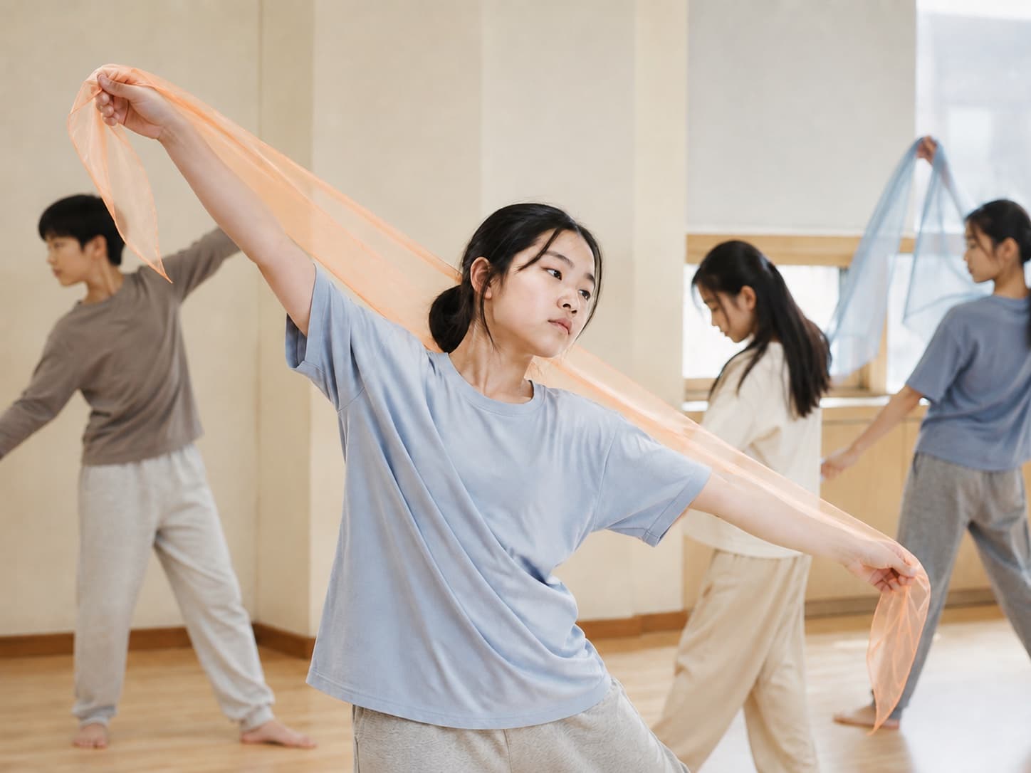 무용실에서 학생들이 얇은 천을 들고 각자 팔을 뻗어 춤추고, 앞의 한 학생이 주황색 천을 펼치고 있다